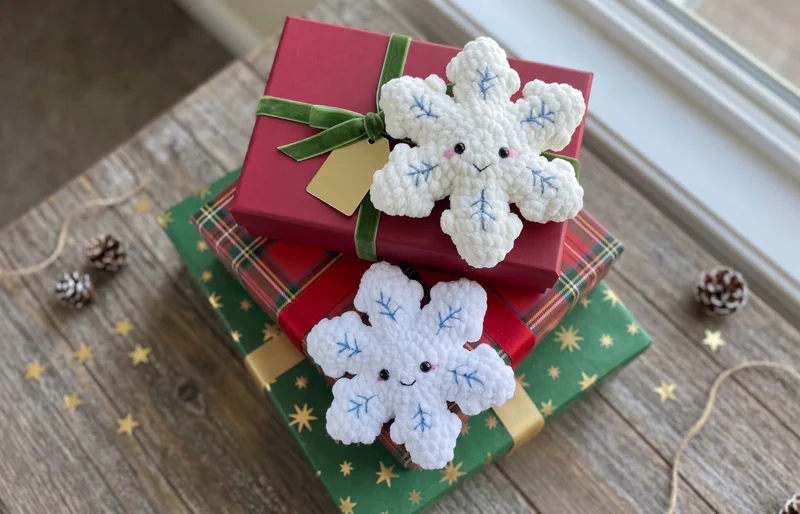 Two smiling white plush crochet snowflakes with blue embroidered branches arranged on wrapped Christmas gifts
