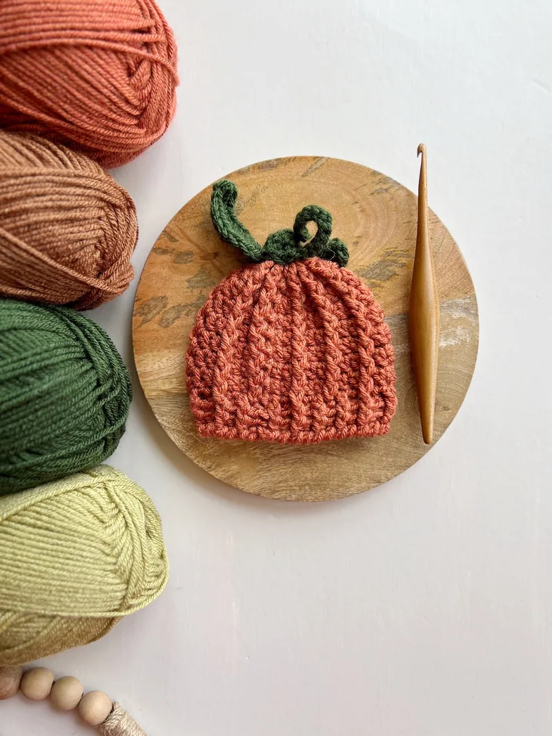 Orange crocheted pumpkin beanie with green stem on a wooden board beside yarn and a crochet hook